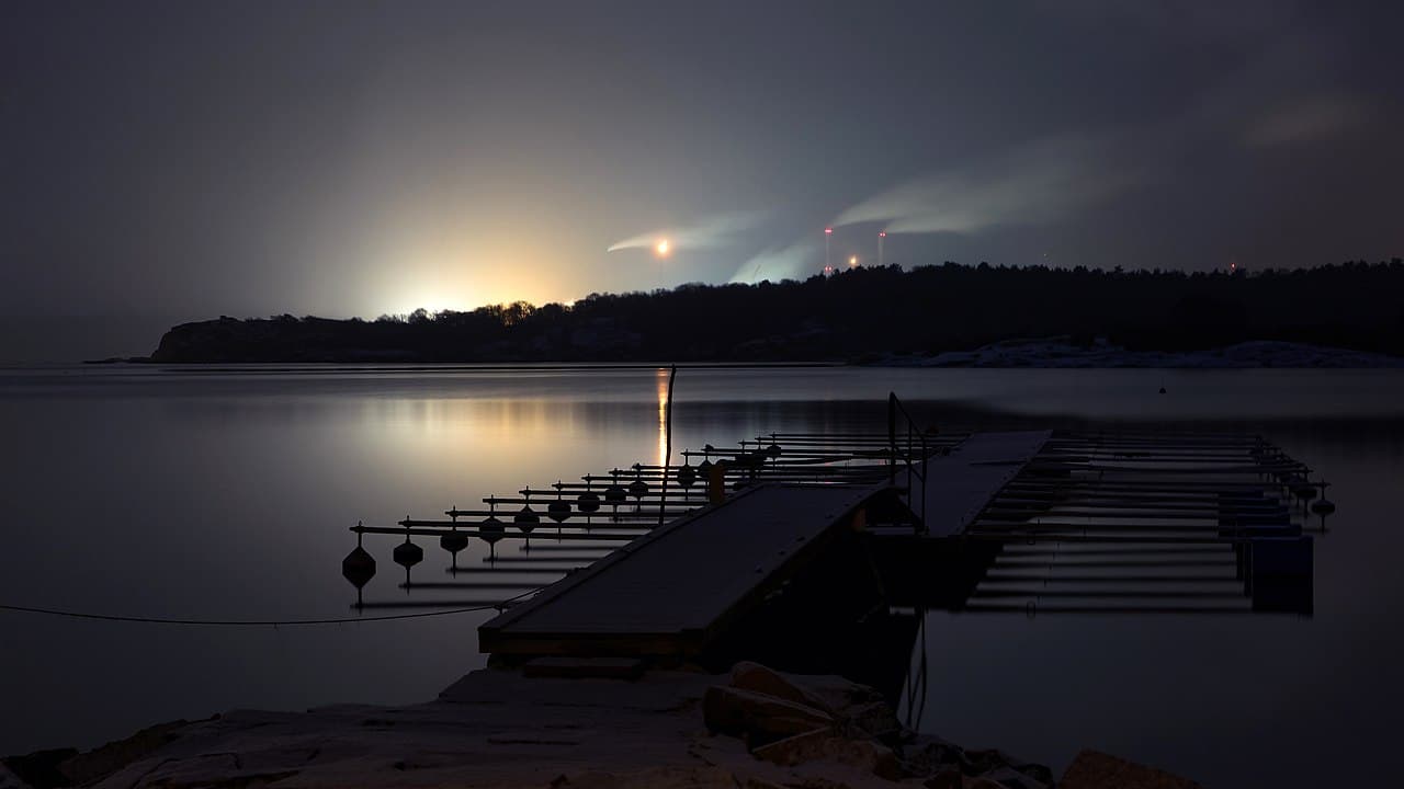 Refinería Preemraff en Brofjorden nevando de noche, con el recinto industrial iluminado en la distancia sobre el fiordo sueco